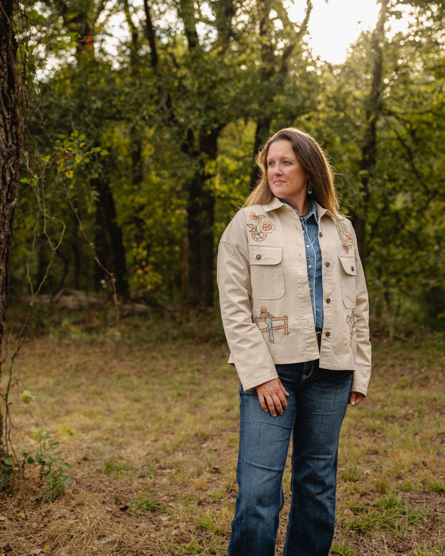 Woman standing in a forest wearing a beige jacket with floral patterns.