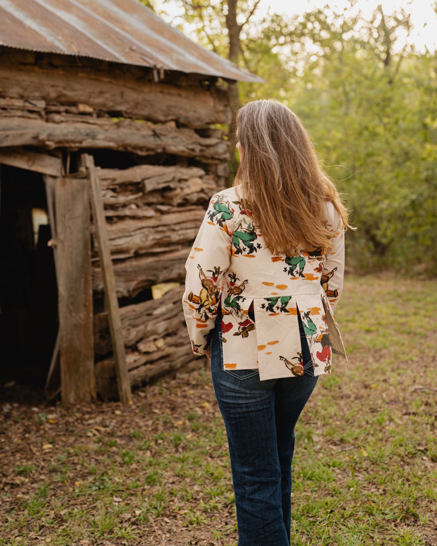 woman walking away wearing jeans and a jacket towards a rustic wooden building with trees in background