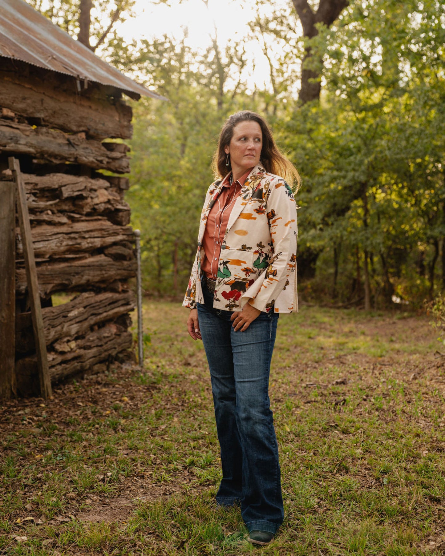 woman wearing jeans and a jacket standing in front of a rustic building with trees in background