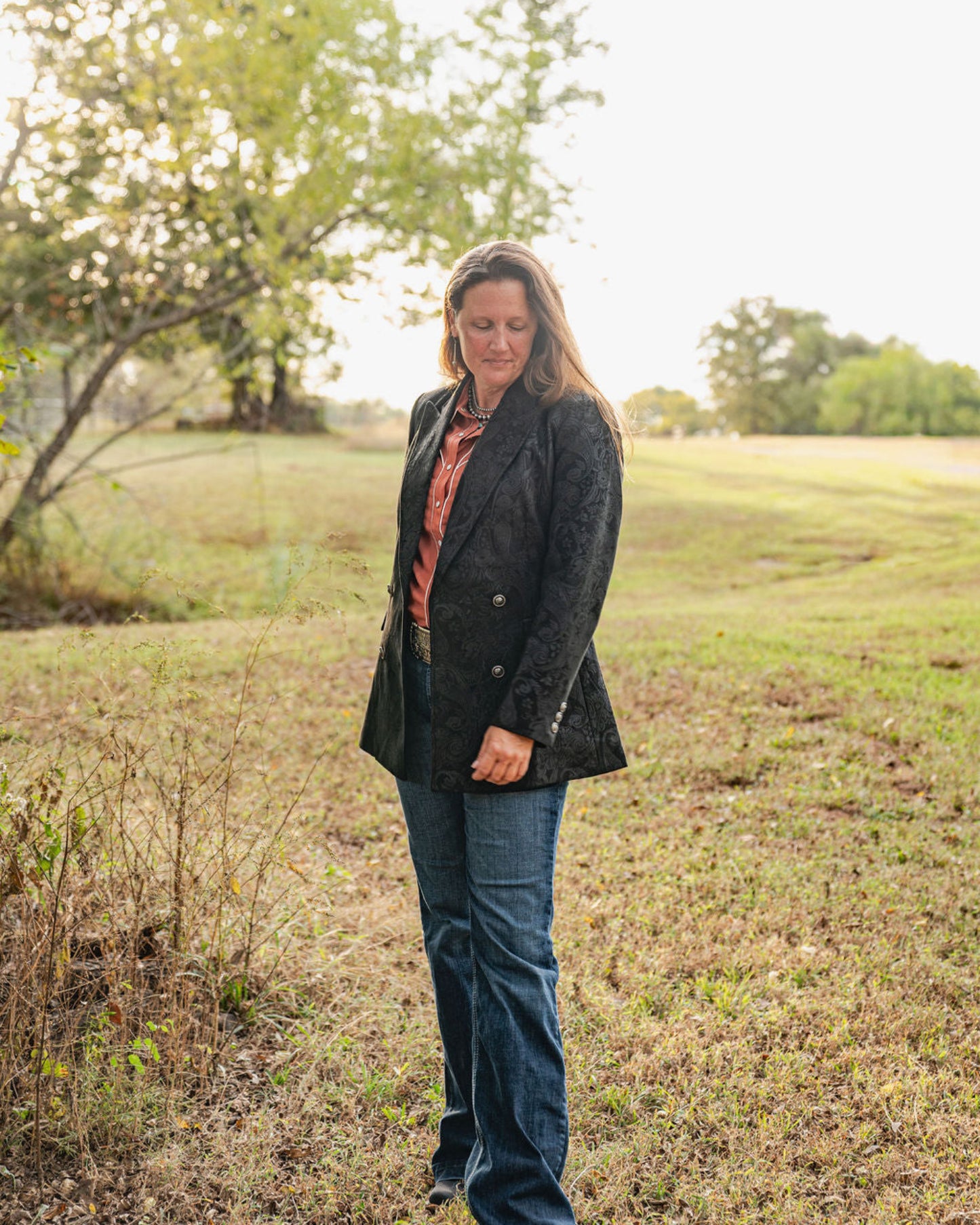 Woman standing in a field wearing a dark coat and blue jeans.