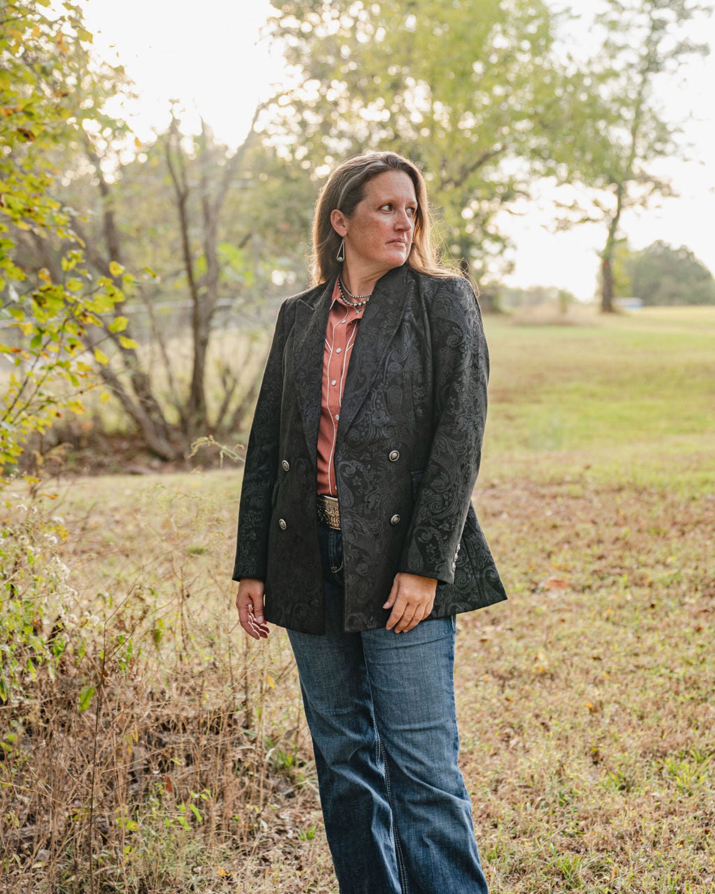 Woman standing in a field wearing a dark coat and jeans.