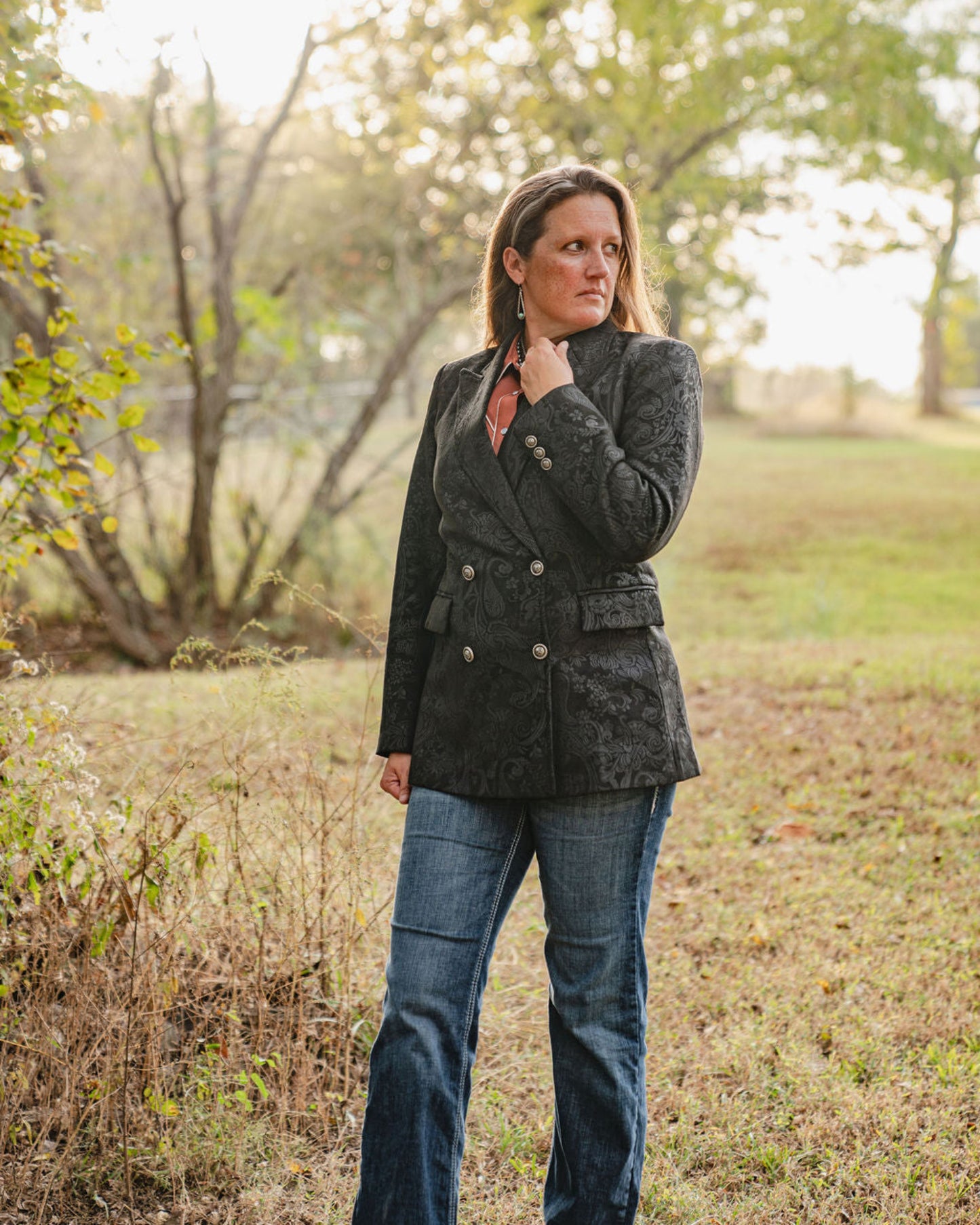 Woman wearing a dark jacket and jeans standing in a field with trees in the background