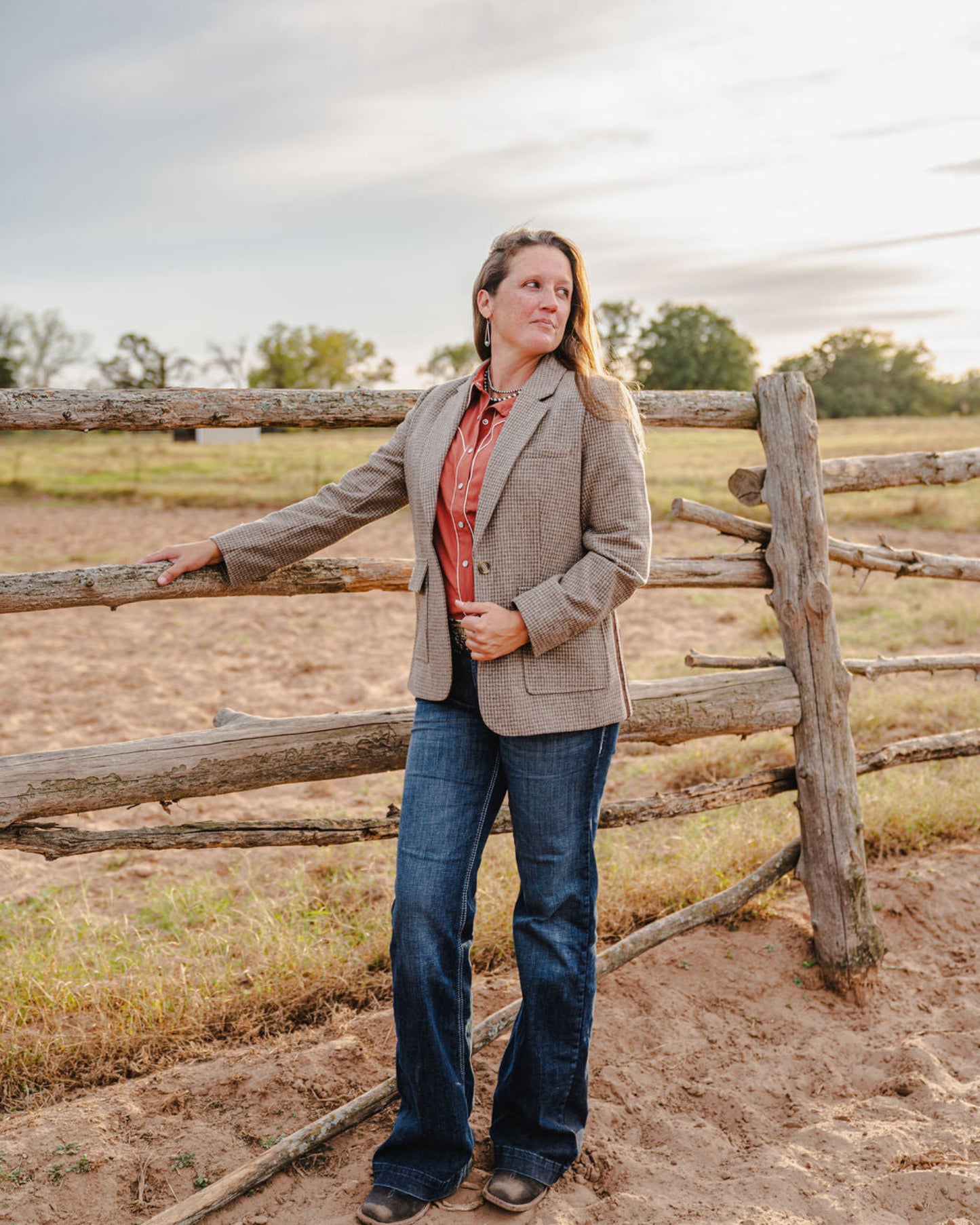 Woman standing by a wooden fence in an open field with a cloudy sky.