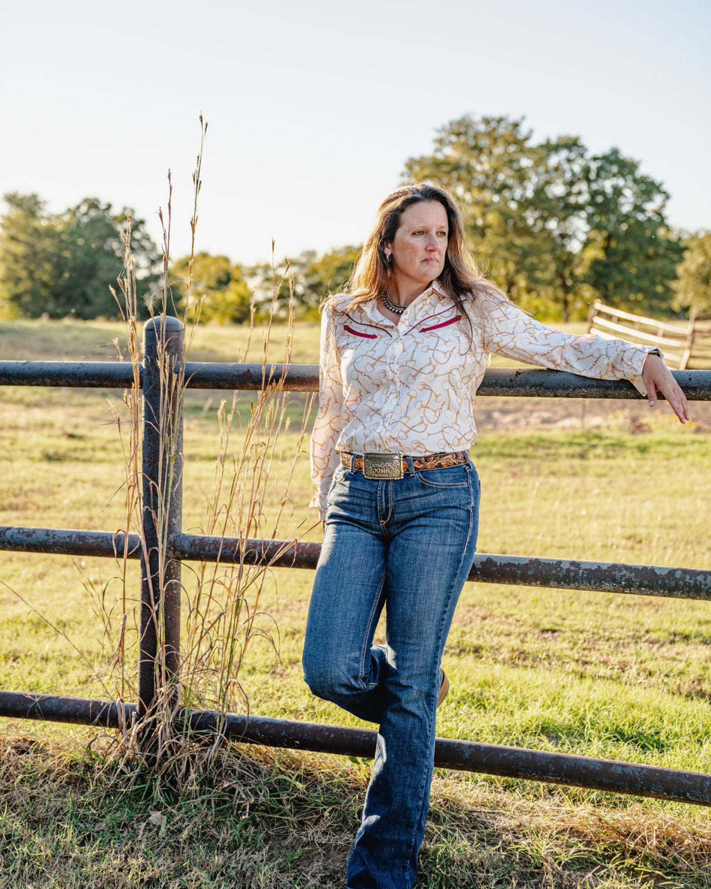 Woman standing by a wooden fence in a grassy field with trees in the background