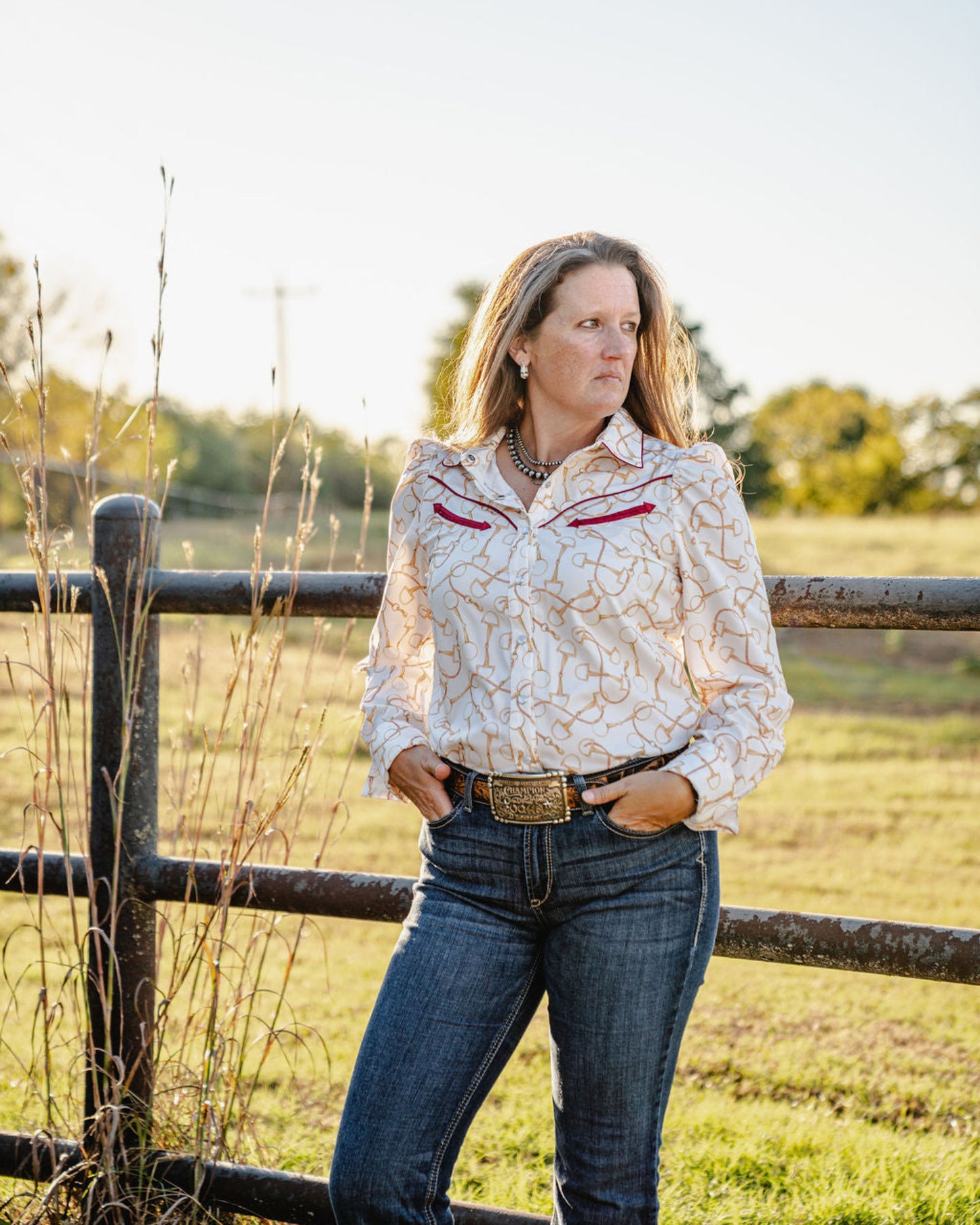 Woman standing in a field wearing a patterned blouse and jeans, with a wooden fence and trees in the background.