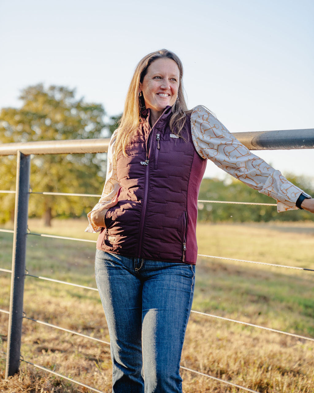 Woman standing in a field wearing a purple vest and jeans, with trees and clear sky in the background.