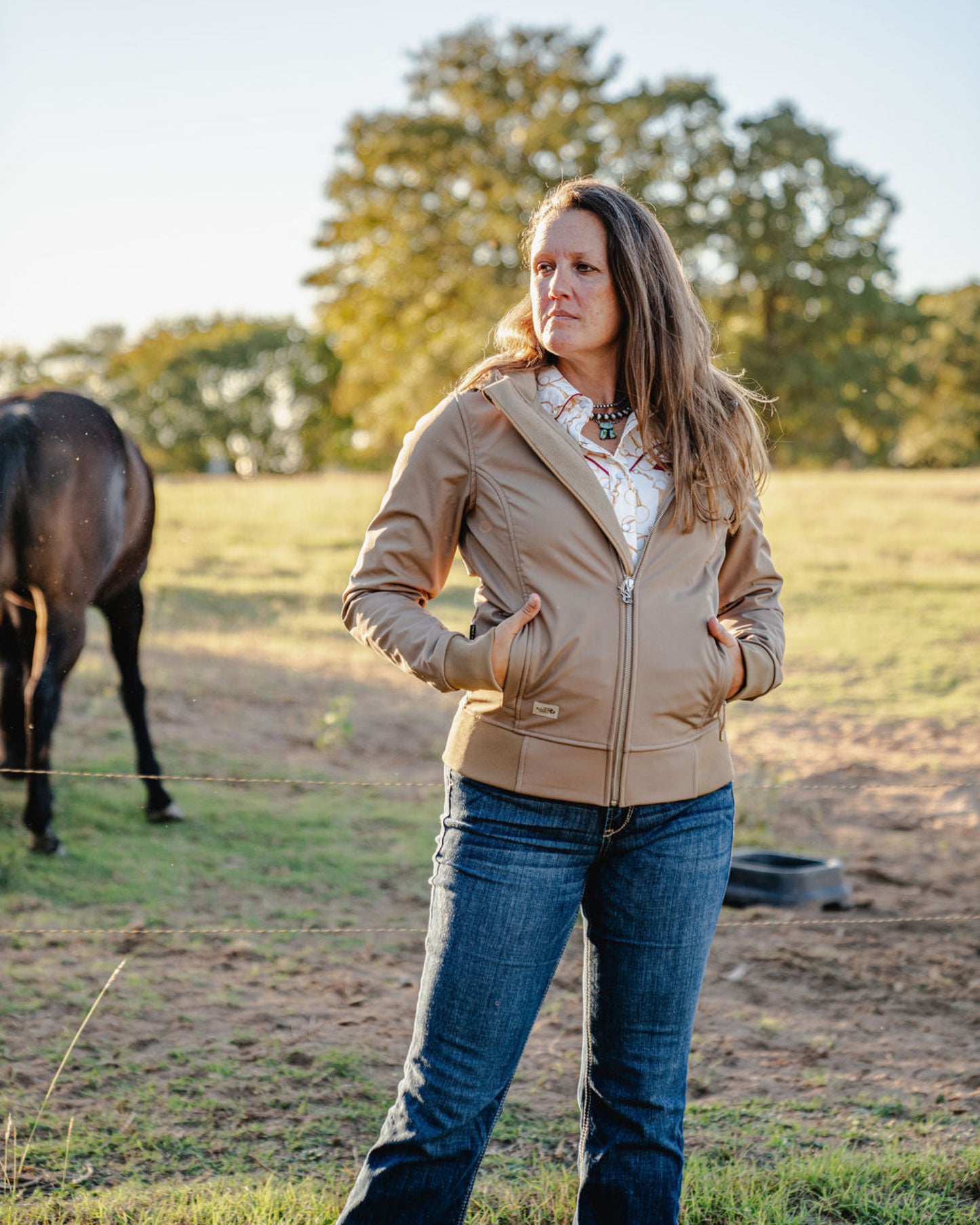 Woman standing in a field with a horse, wearing a beige jacket and blue jeans.