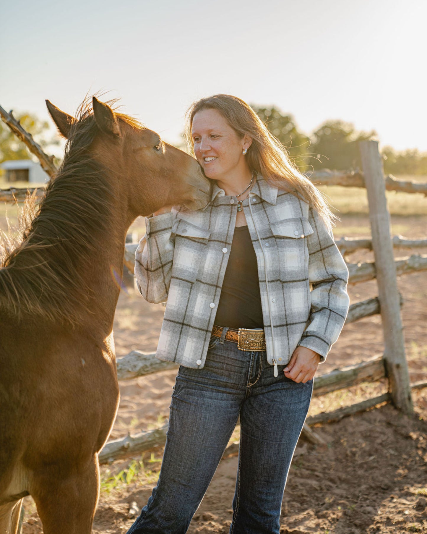 Woman standing next to a horse in an outdoor setting with a wooden fence and trees in the background.