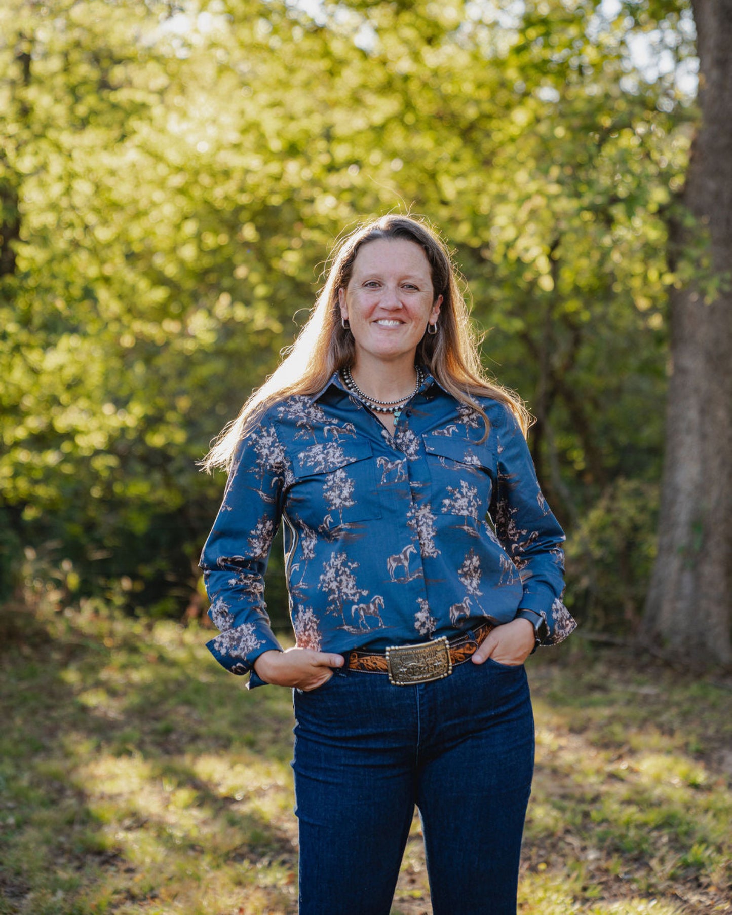 Woman wearing a blue floral shirt and jeans standing in a park with trees in the background