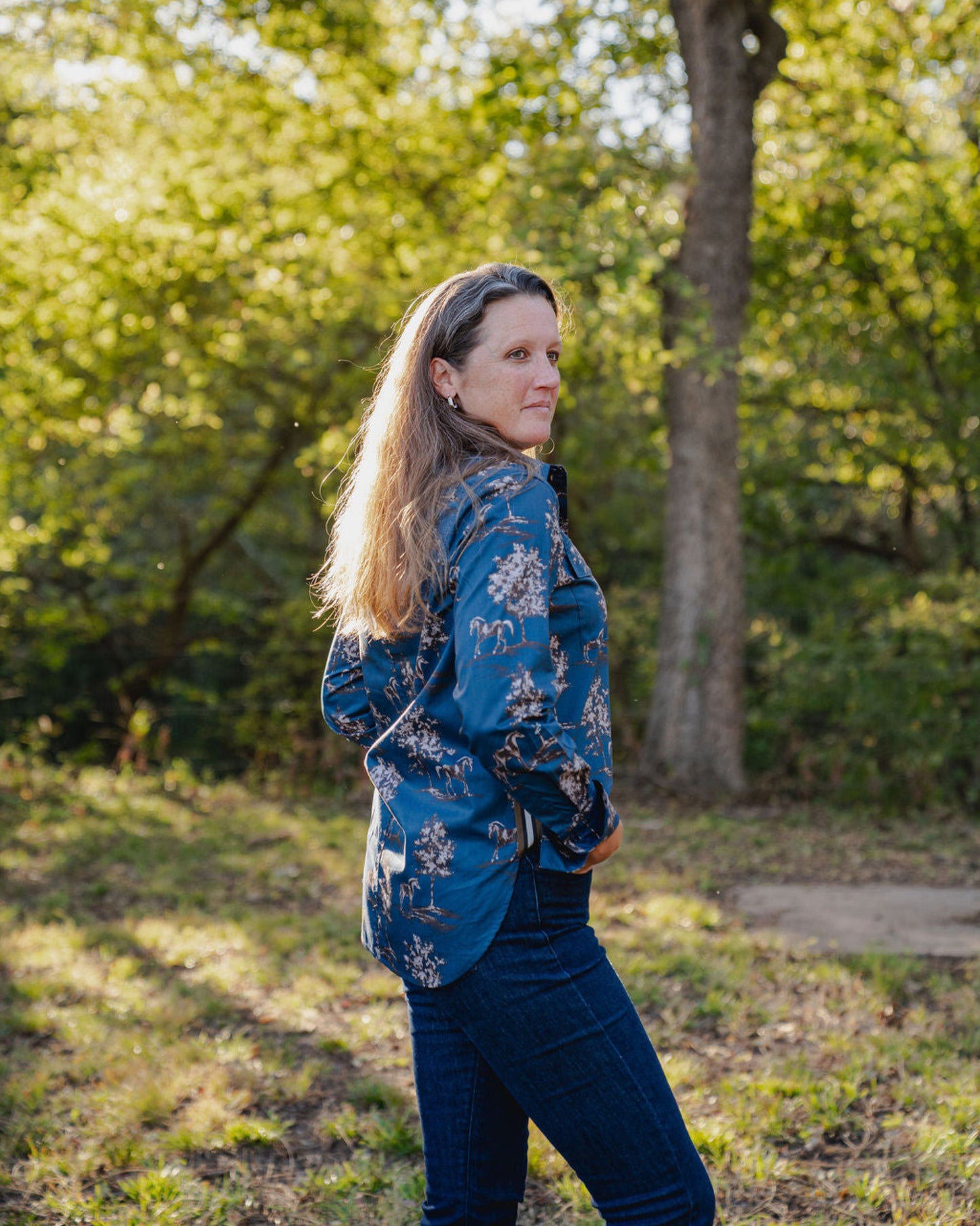 Woman wearing a blue floral jacket and dark pants standing in a park with trees in the background