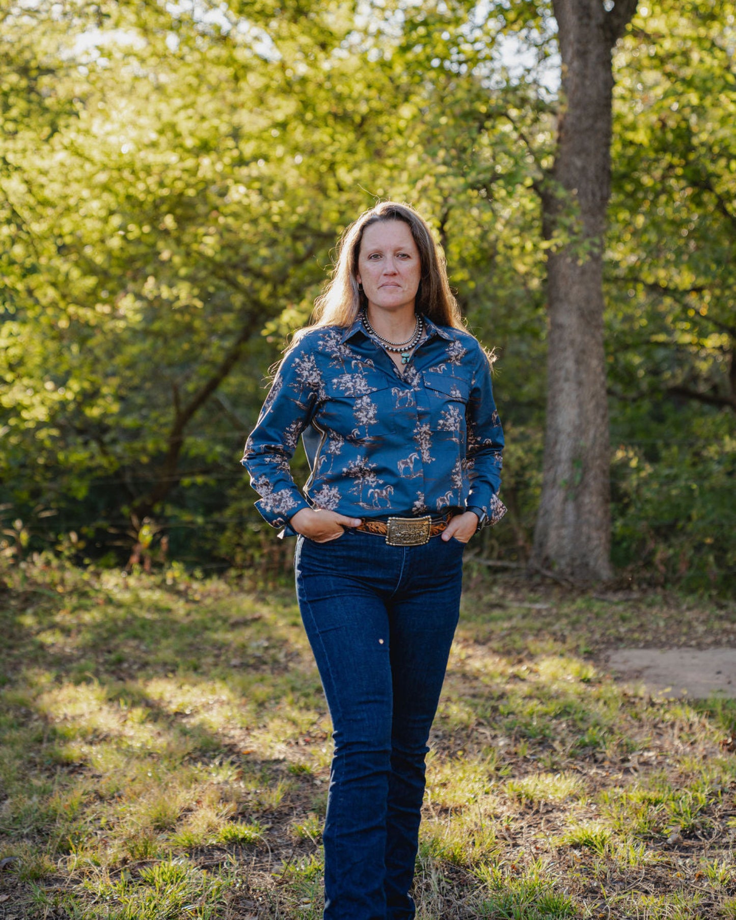 Woman wearing a blue floral shirt and jeans standing in a grassy area with trees.