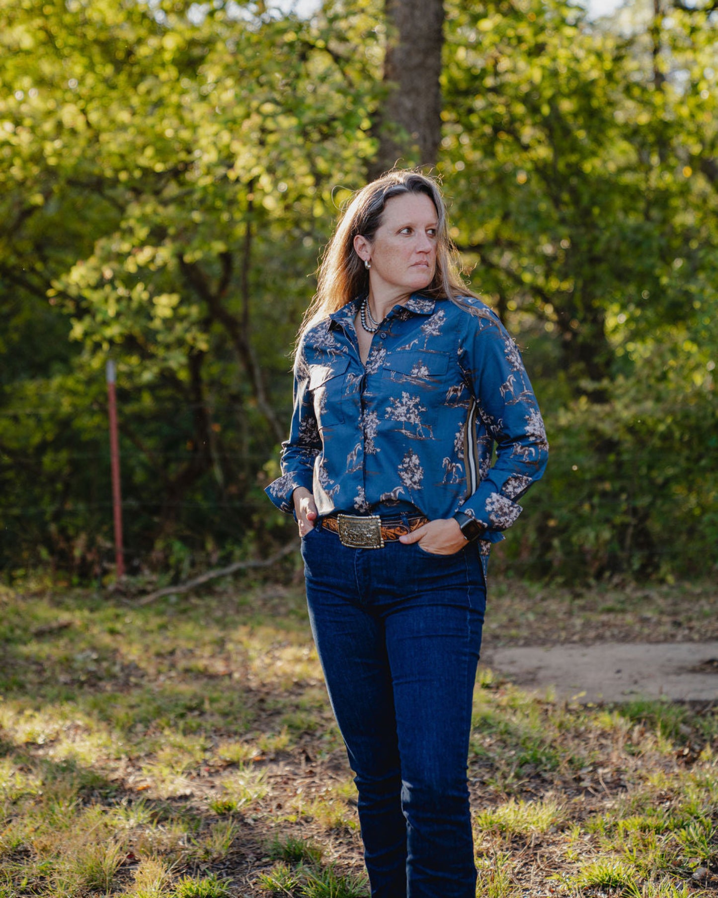Woman wearing a blue floral shirt and jeans standing in a forested area.