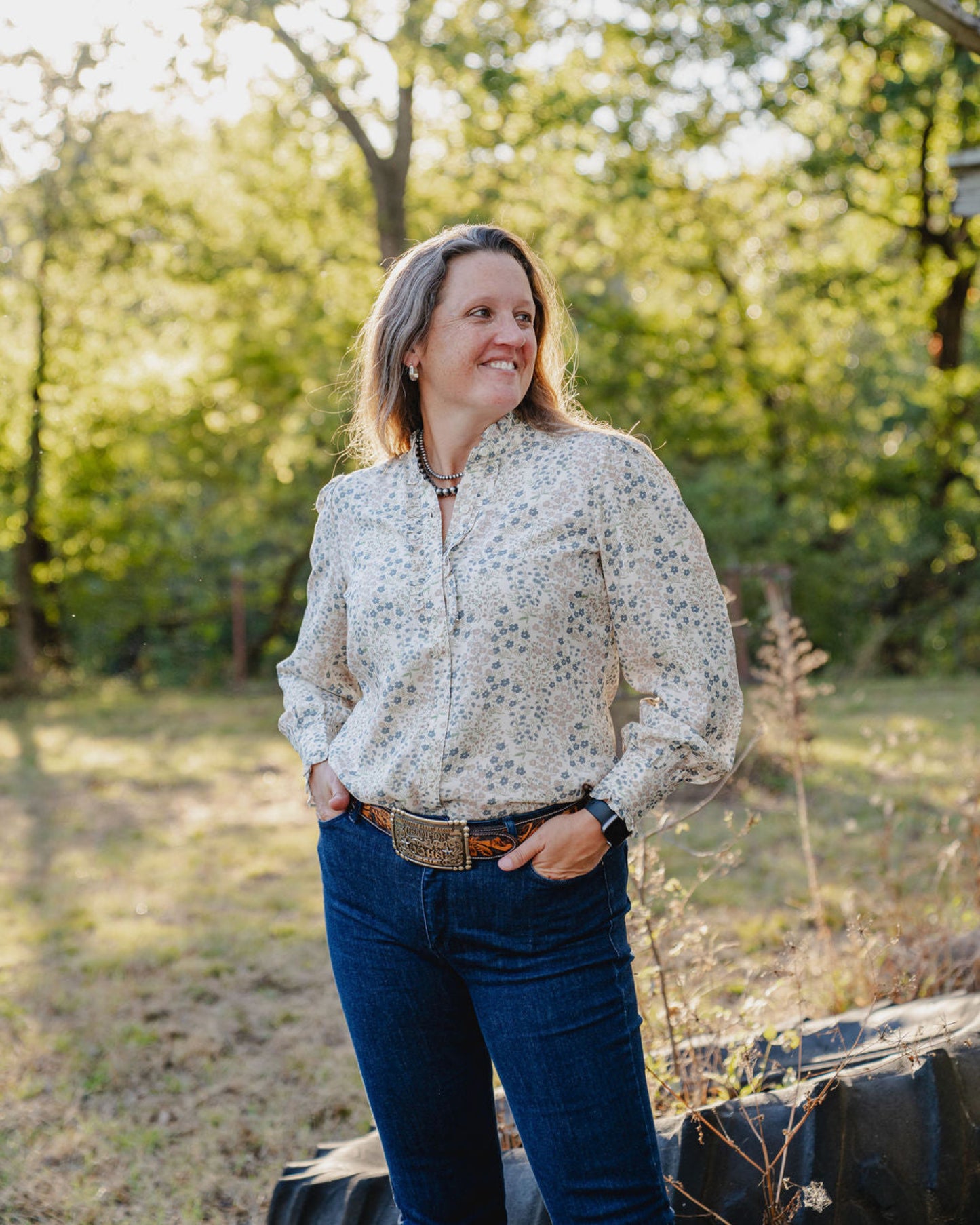 Woman standing outdoors in a natural setting with trees and greenery.
