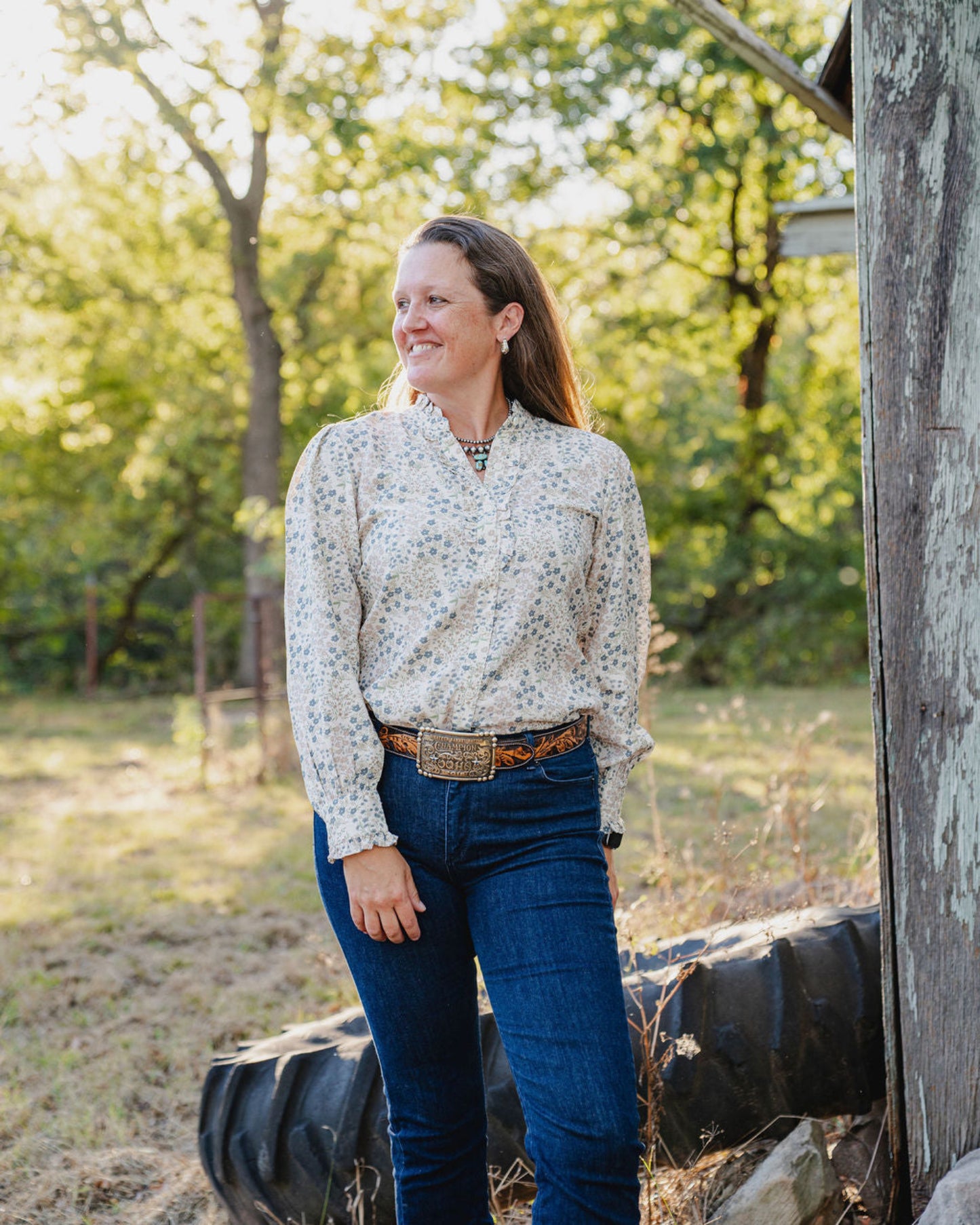 Woman standing outdoors in a natural setting with trees and a wooden structure.