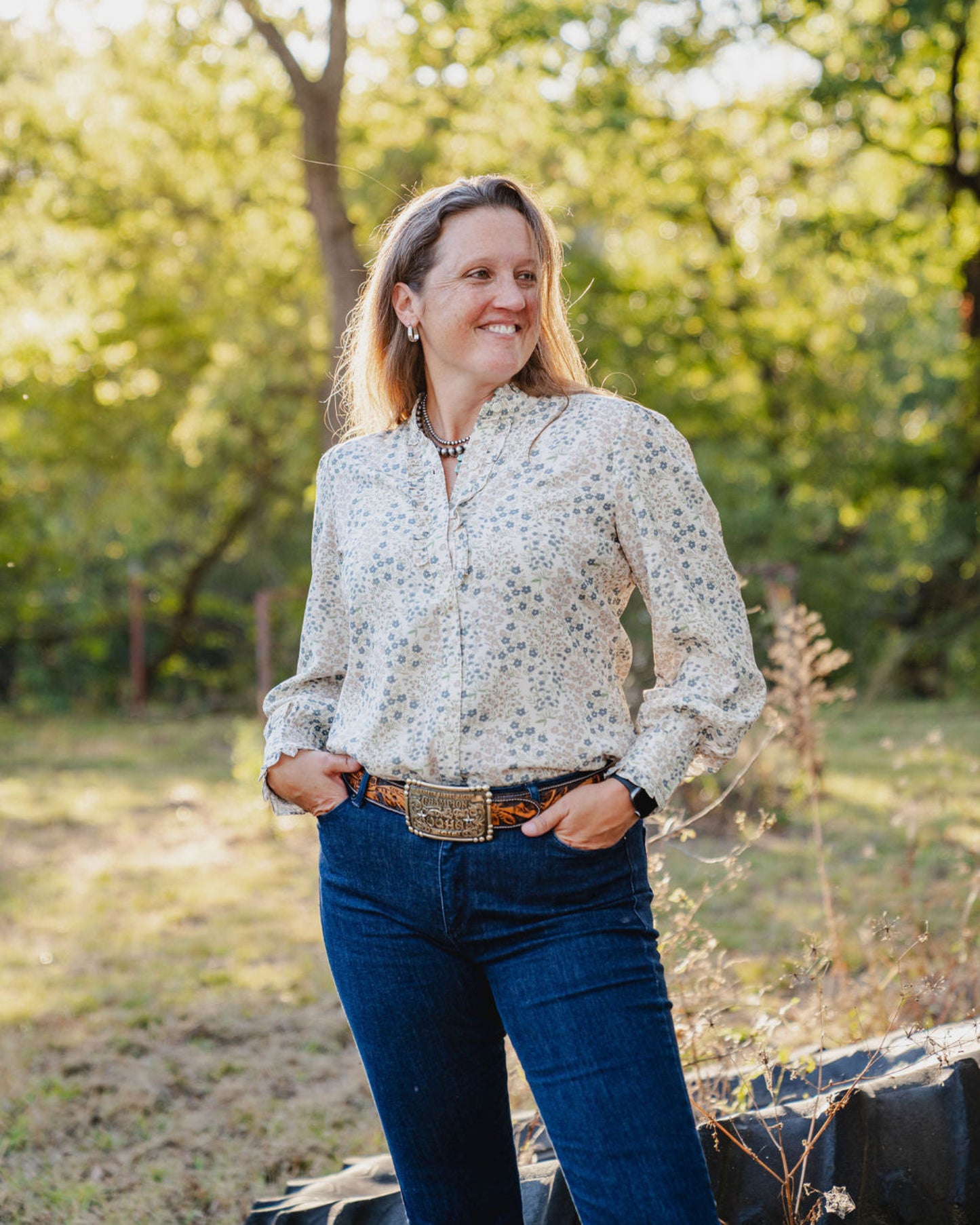 Woman standing outdoors in a natural setting with trees and greenery.