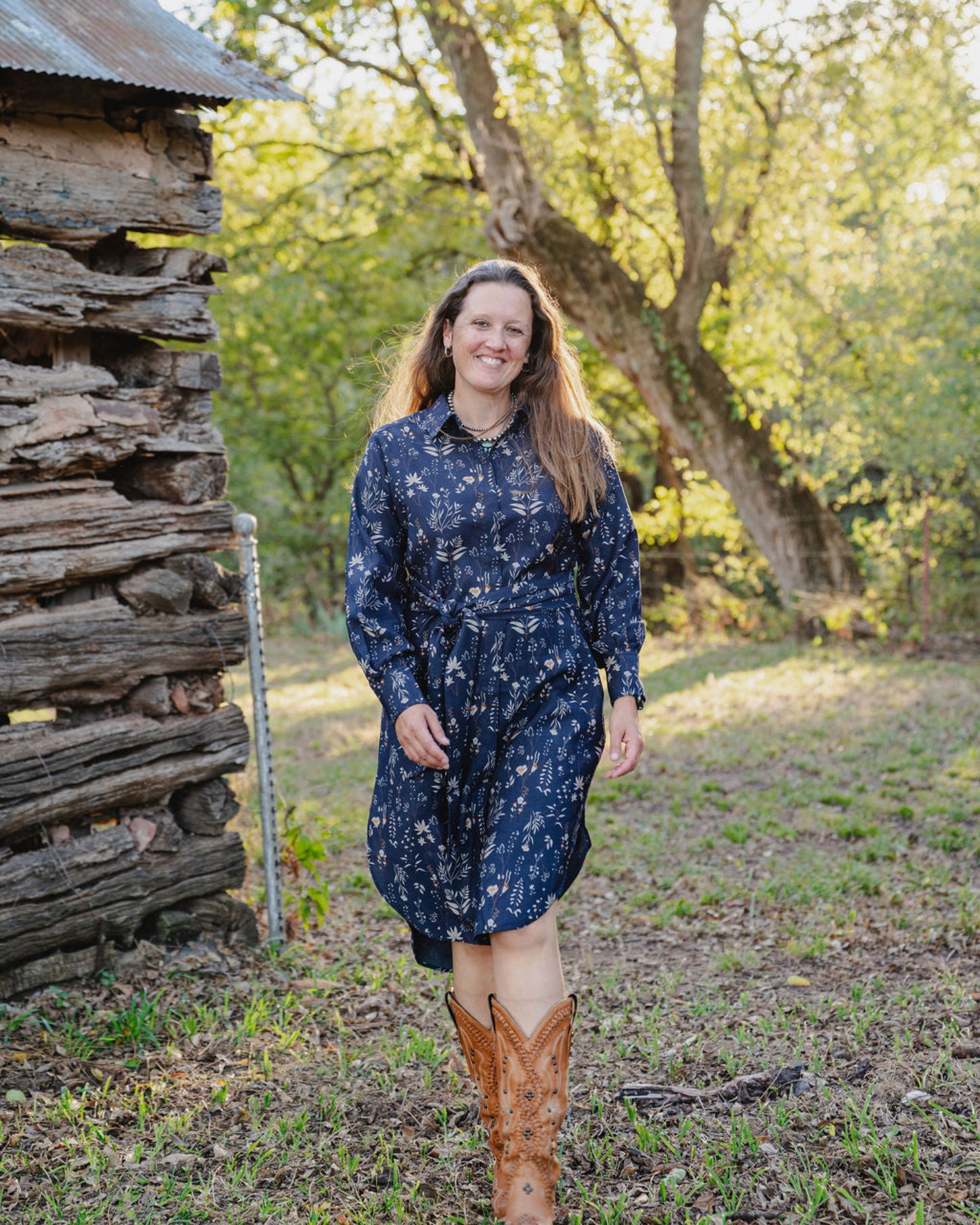 Woman in a blue dress standing in a natural setting with trees and a wooden structure.
