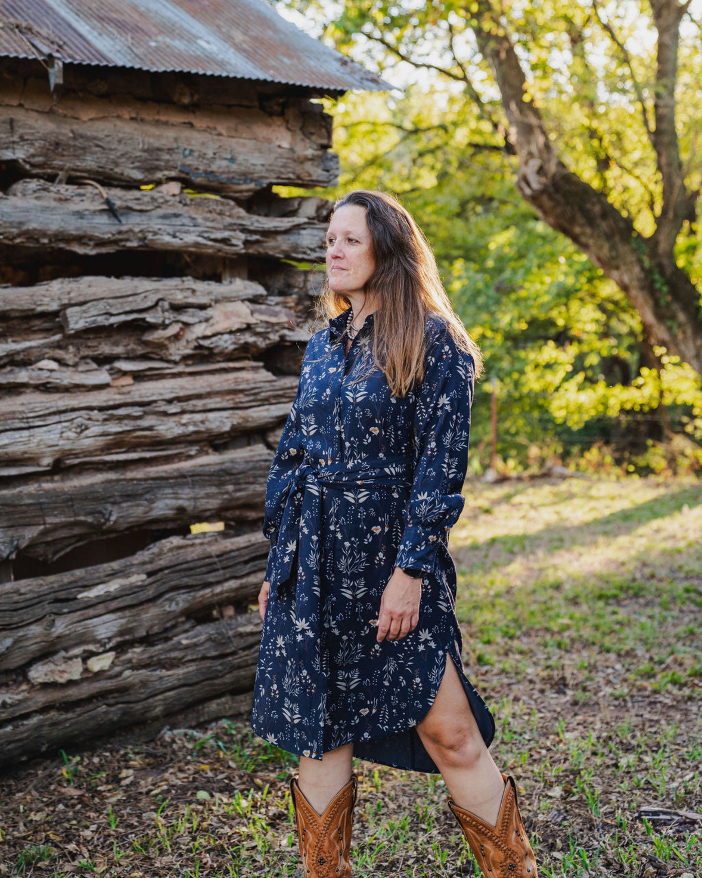 Woman in a blue floral dress standing in front of a rustic wooden cabin.