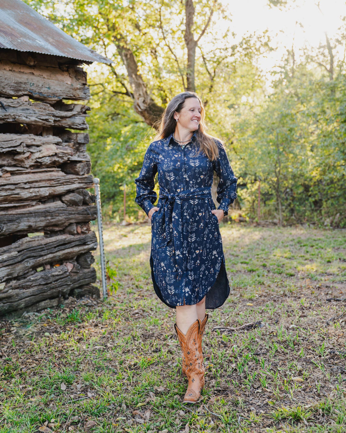 Woman in a blue floral dress and brown boots standing in a natural setting with trees and a wooden structure.