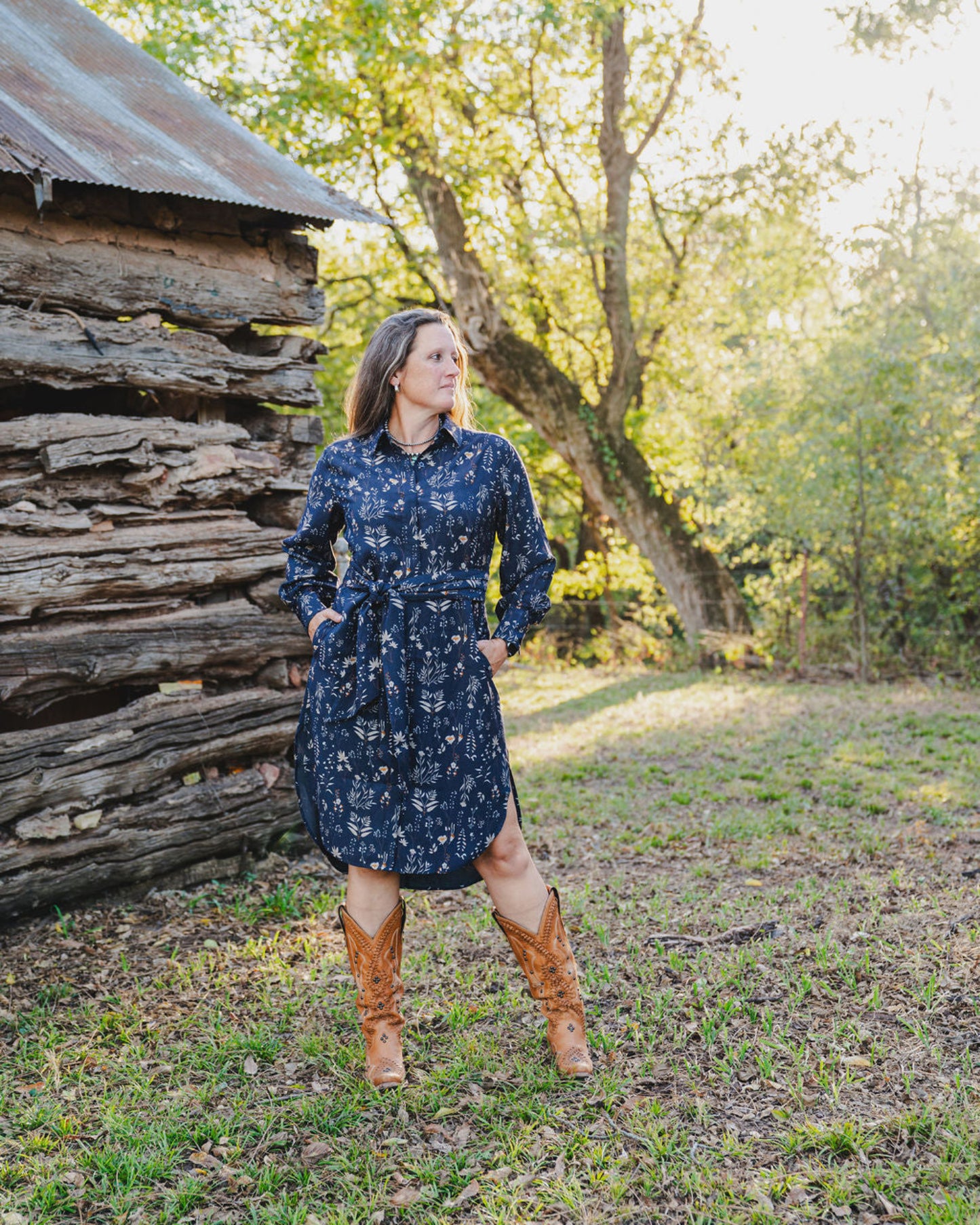 Woman in a blue floral dress and brown boots standing in a natural setting with trees and wooden logs.