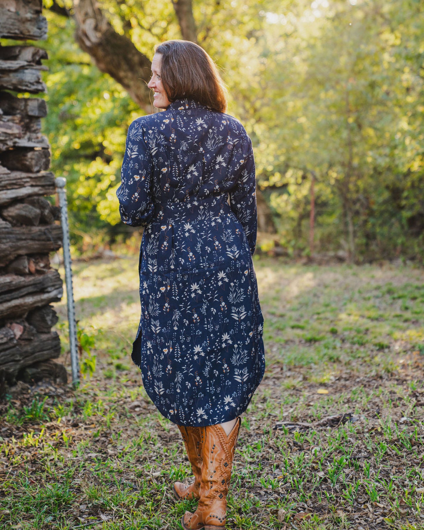Woman in a navy floral dress standing in a forested area with a wooden structure.