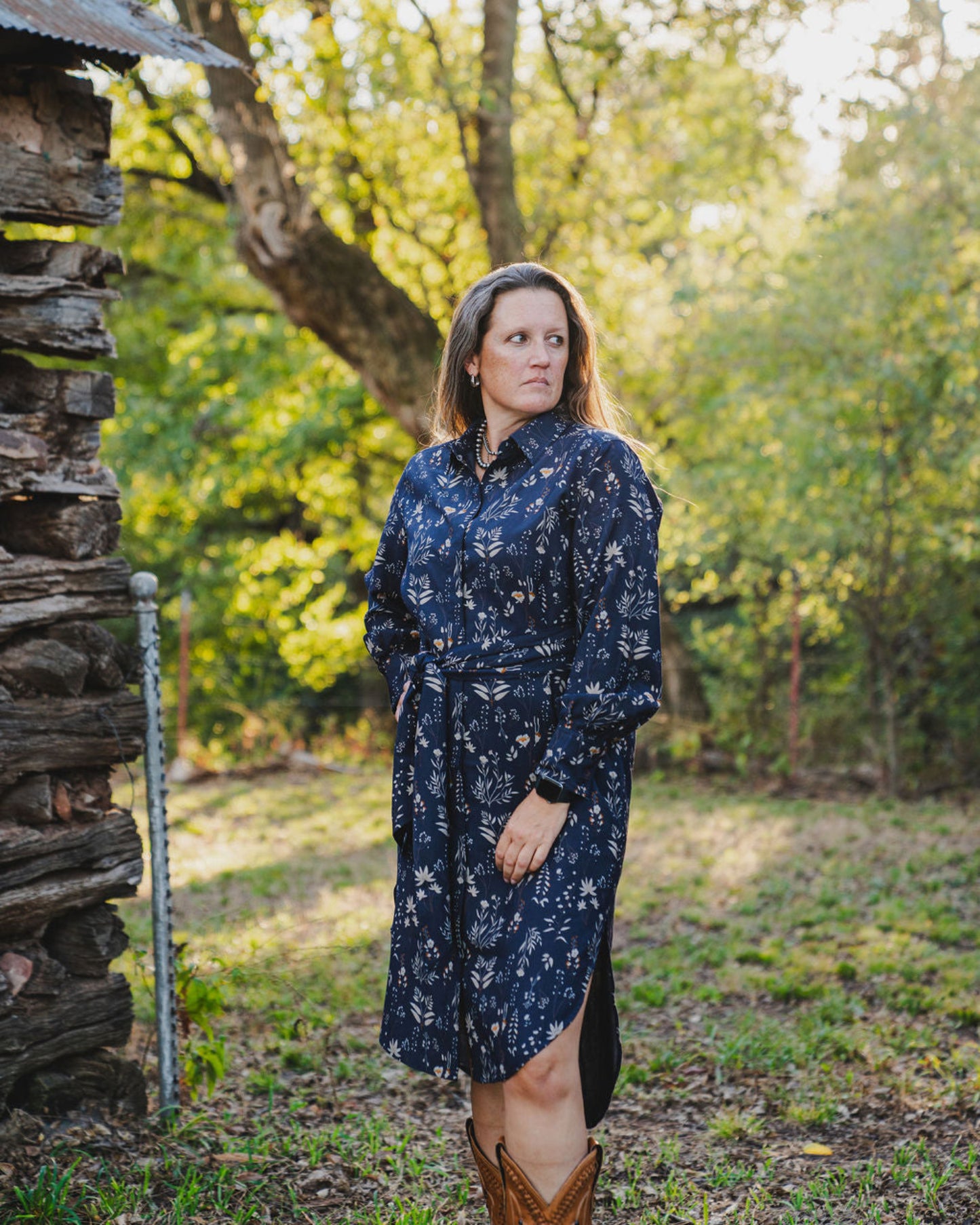 Woman in a blue floral dress standing in a natural setting with trees and a wooden structure.