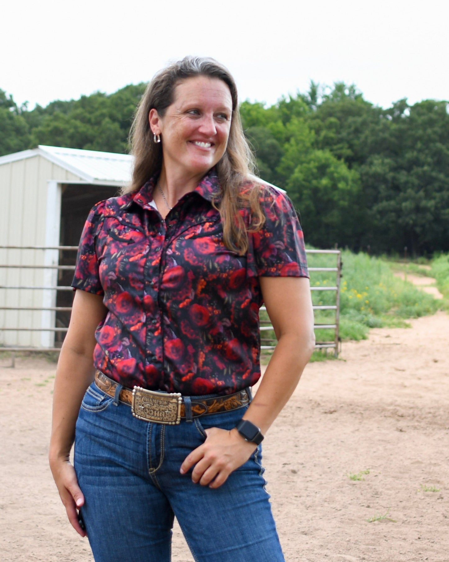 Woman wearing a patterned shirt and jeans standing outdoors with trees and a building in the background