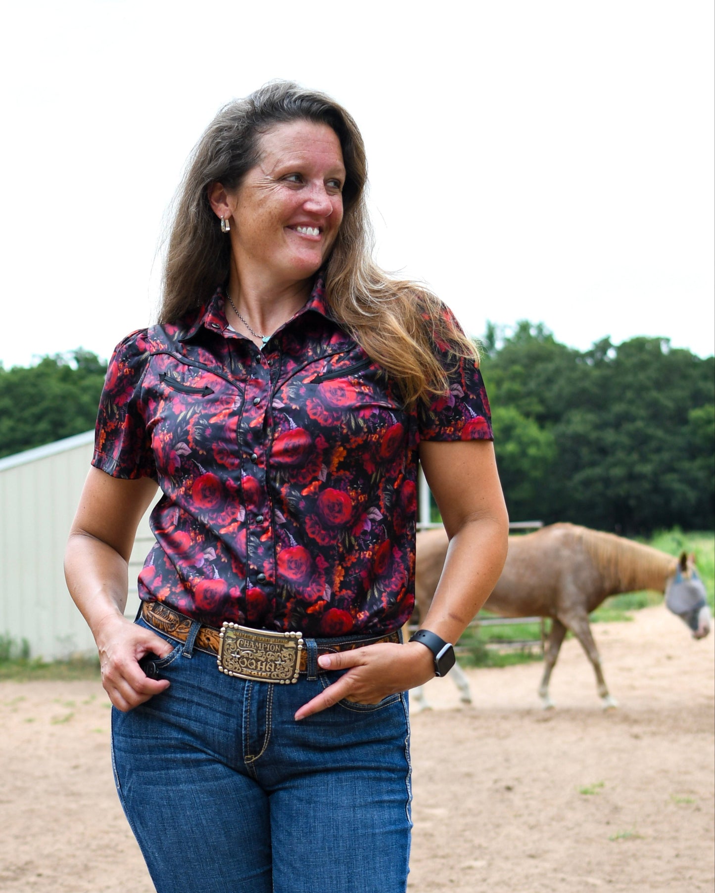 Woman in a patterned shirt and jeans standing in an outdoor setting with a horse in the background