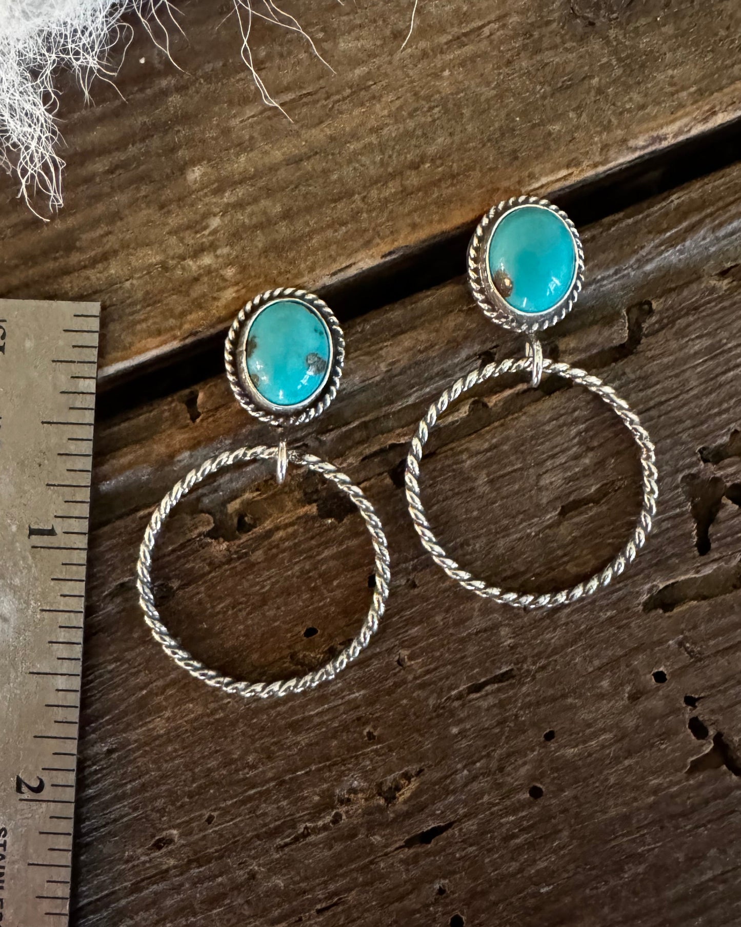 A pair of silver-toned dangle earrings with oval turquoise-blue stones and twisted hoop drops, displayed on a rustic wooden surface next to a metal ruler for scale.