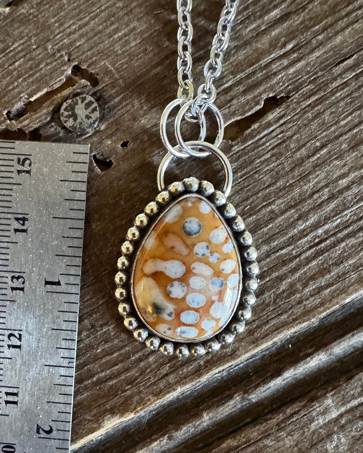 A silver beaded pendant necklace with a teardrop-shaped orange and white patterned stone, displayed next to a metal ruler on a rustic wooden surface.