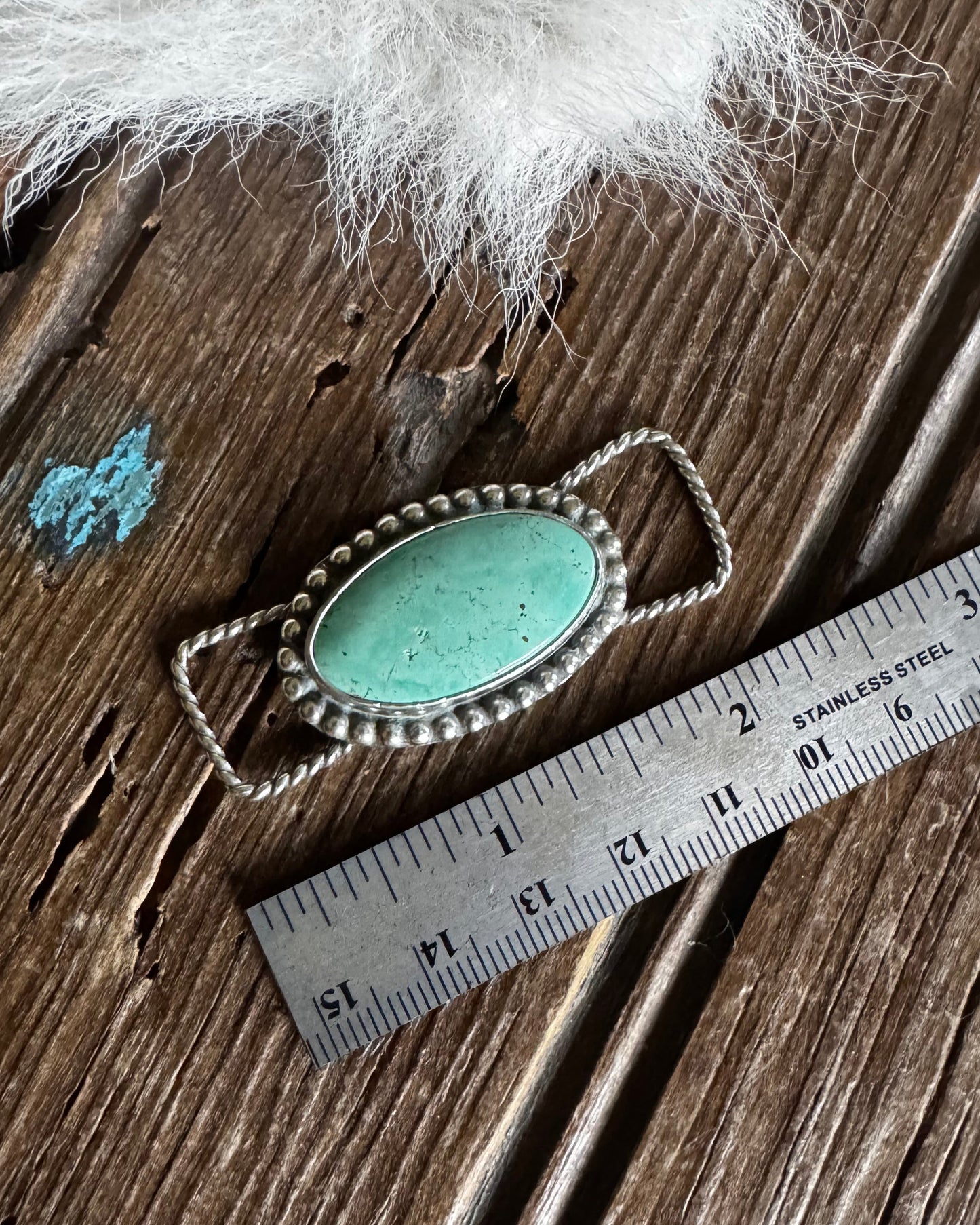 Turquoise pendant with silver frame on a wooden surface next to a ruler