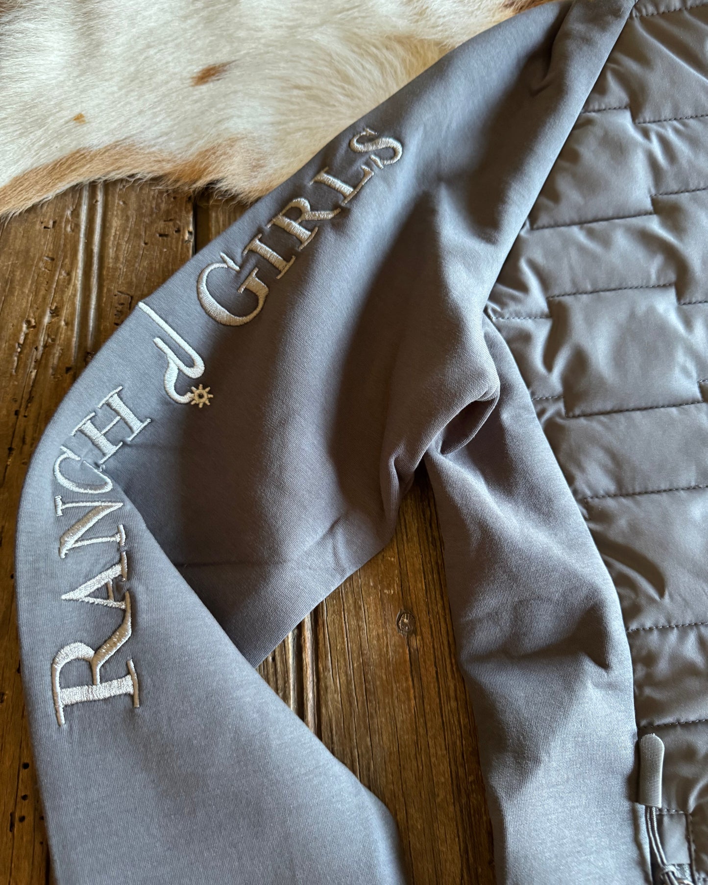 Gray quilted jacket with 'Ranch Girls' embroidery on a wooden surface.