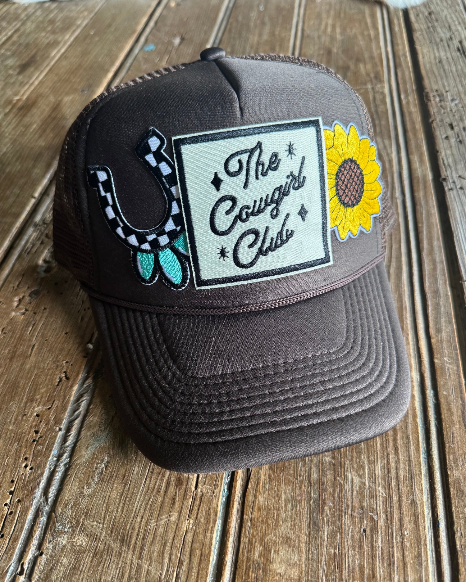 brown foam trucker with sunflower design and 'The Girl Cowgirl Club' text on a wooden surface.