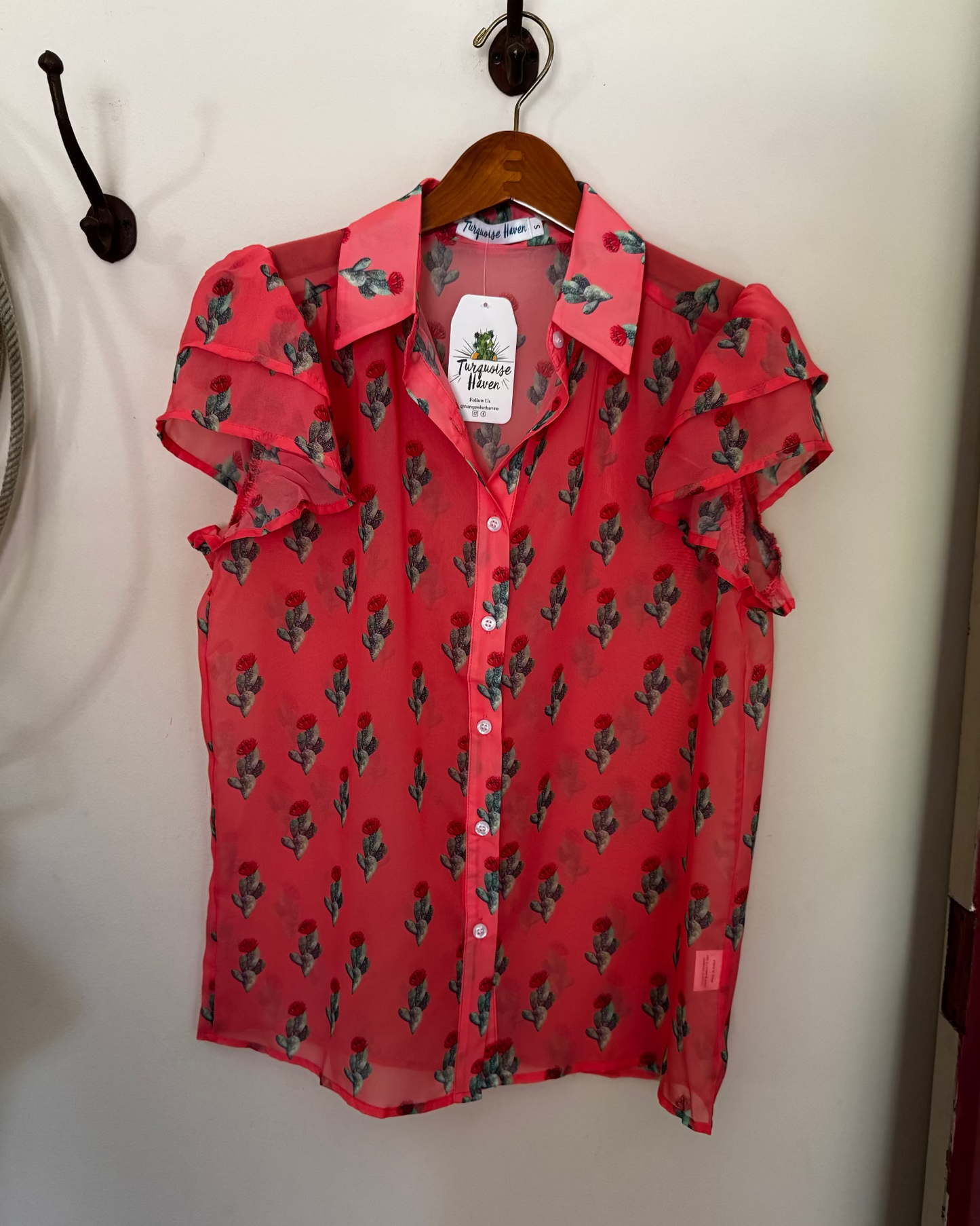 A red short-sleeve button-up blouse with a green cactus print, hanging on a wooden hanger against a white wall.