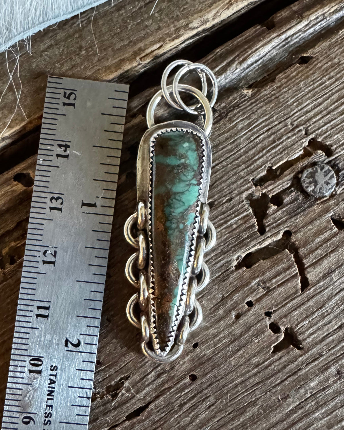 A handmade sterling silver pendant with a green turquoise stone, shown next to a stainless steel ruler on a rustic wooden surface.