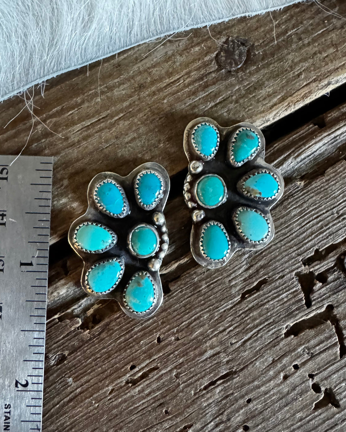 Two silver-toned floral turquoise stud earrings displayed on a rustic wooden surface next to a stainless steel ruler for scale.