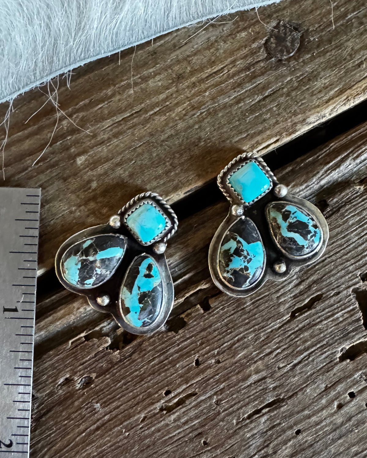 A pair of turquoise and stone drop earrings displayed on a rustic wooden surface next to a metal ruler for scale.
