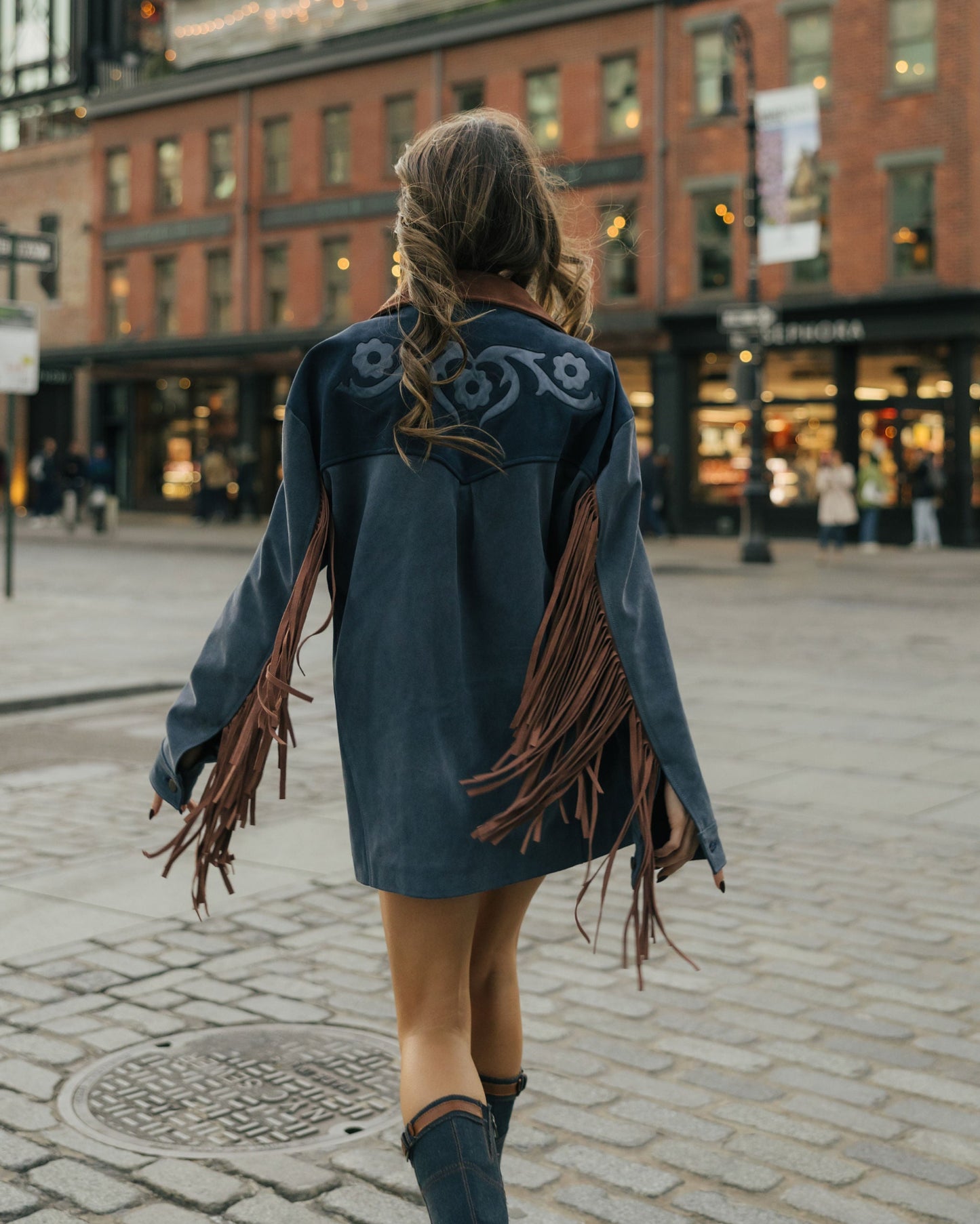Person wearing a blue leather jacket with fringe walking on a city street.
