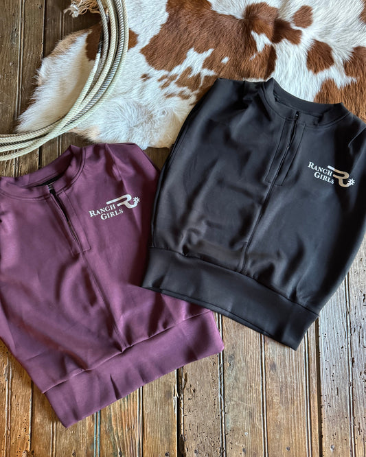 Black and purple shirts with 'Ranch Girl' branding on a wooden surface with cowhide rug.