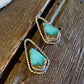 A pair of gold-toned drop earrings with triangular turquoise-blue stones, resting on a rustic dark wood surface next to a white faux-fur texture.