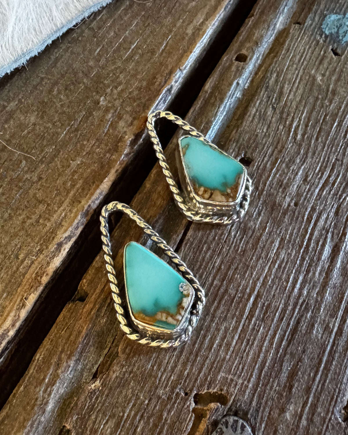 A pair of gold-toned drop earrings with triangular turquoise-blue stones, resting on a rustic dark wood surface next to a white faux-fur texture.