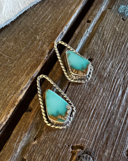 A pair of gold-toned drop earrings with triangular turquoise-blue stones, resting on a rustic dark wood surface next to a white faux-fur texture.
