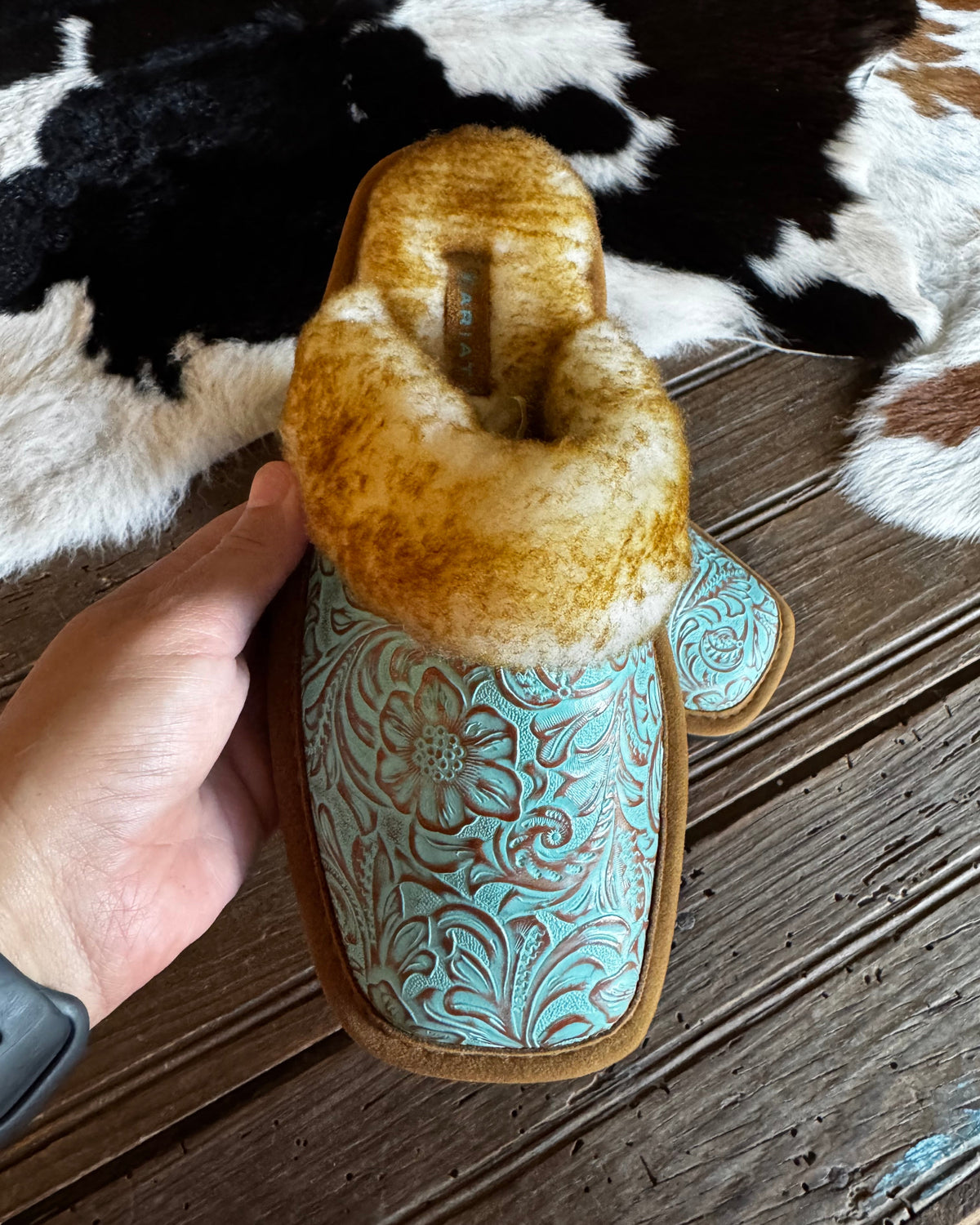 hand holding a turquoise embossed leather slipper above a wooden and cowhide background