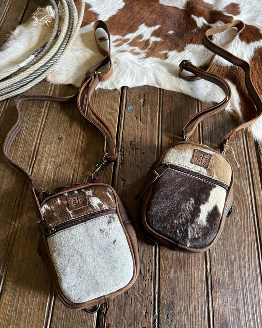 Two cowhide-patterned bags on a wooden surface with a cowhide rug and lasso.