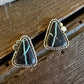A pair of triangular turquoise and black stone stud earrings with silver-toned bezel settings, resting on a rustic wooden surface next to a white faux fur trim.