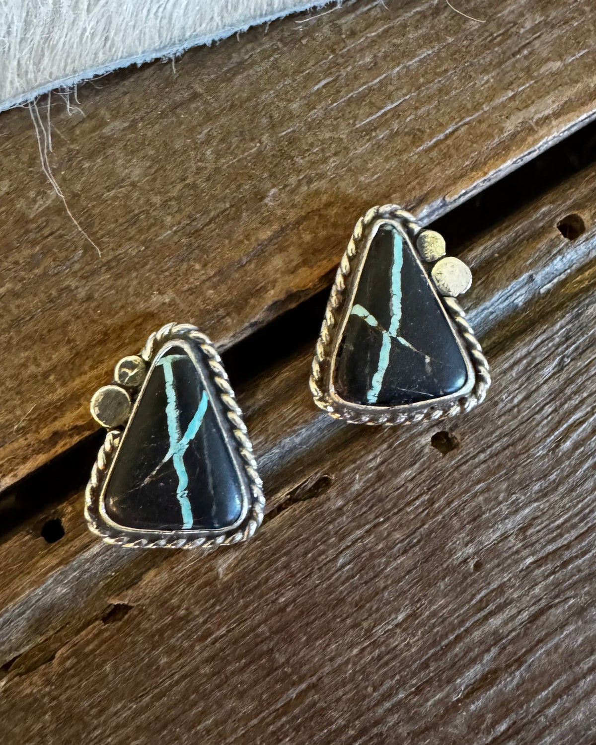 A pair of triangular turquoise and black stone stud earrings with silver-toned bezel settings, resting on a rustic wooden surface next to a white faux fur trim.