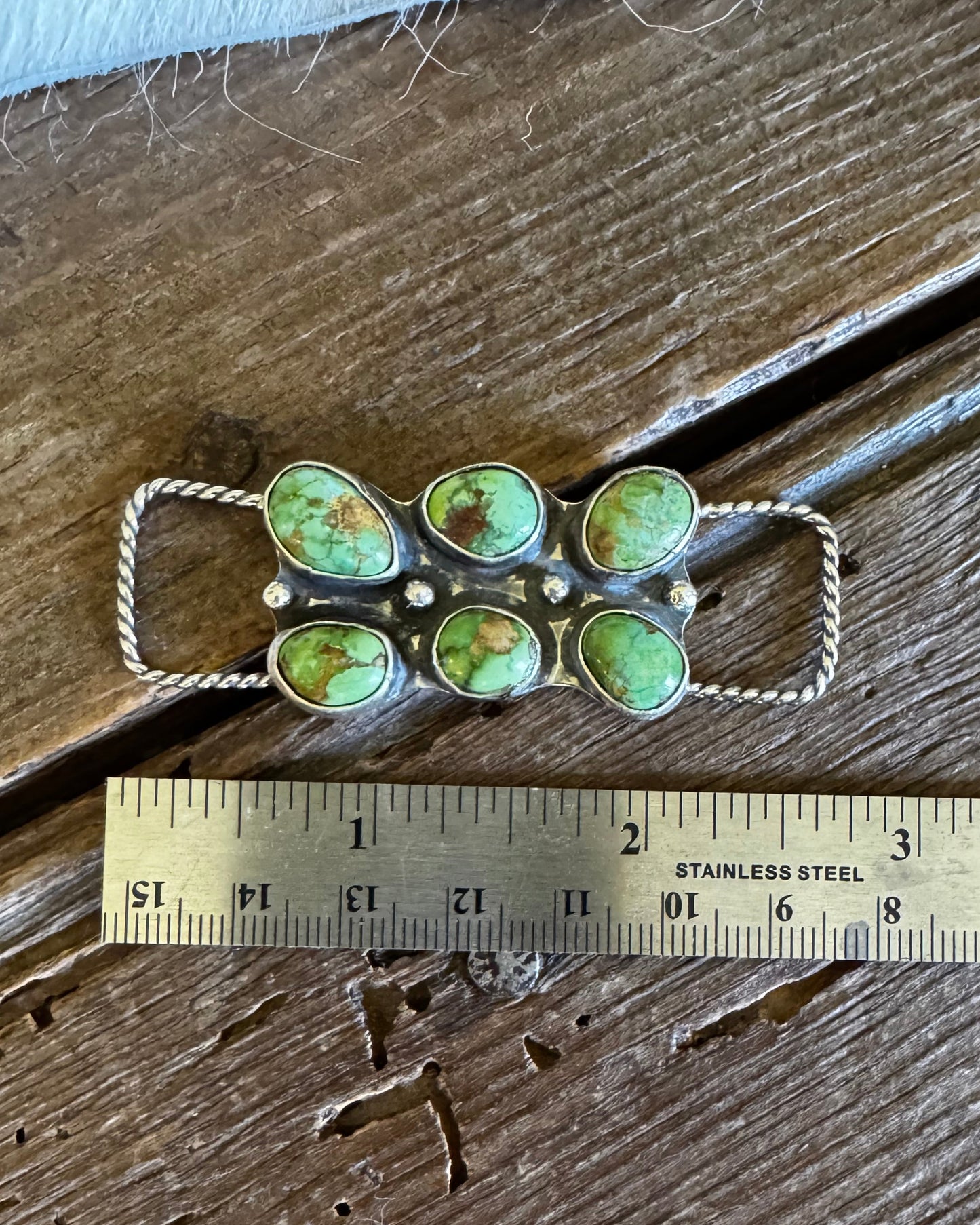 Jewelry pendant with green stones and silver accents on a wooden surface with a ruler for scale.