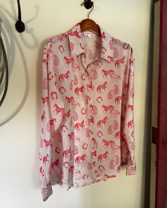 A long-sleeve button-up shirt with a pink and red horse pattern, hanging on a wooden hanger from a black wall hook.