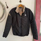Black puffer jacket with fur lining hanging on a hook against a white wall.