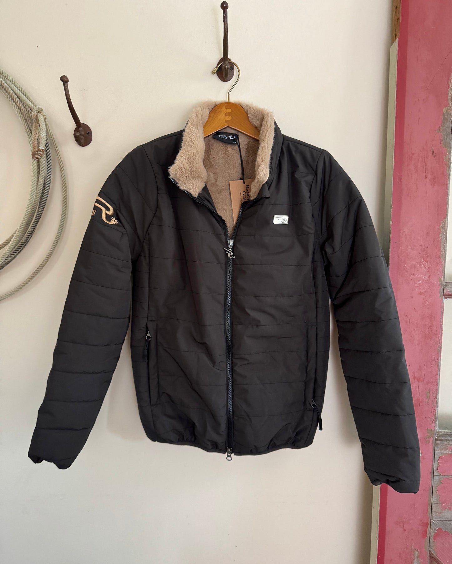 Black puffer jacket with fur lining hanging on a hook against a white wall.