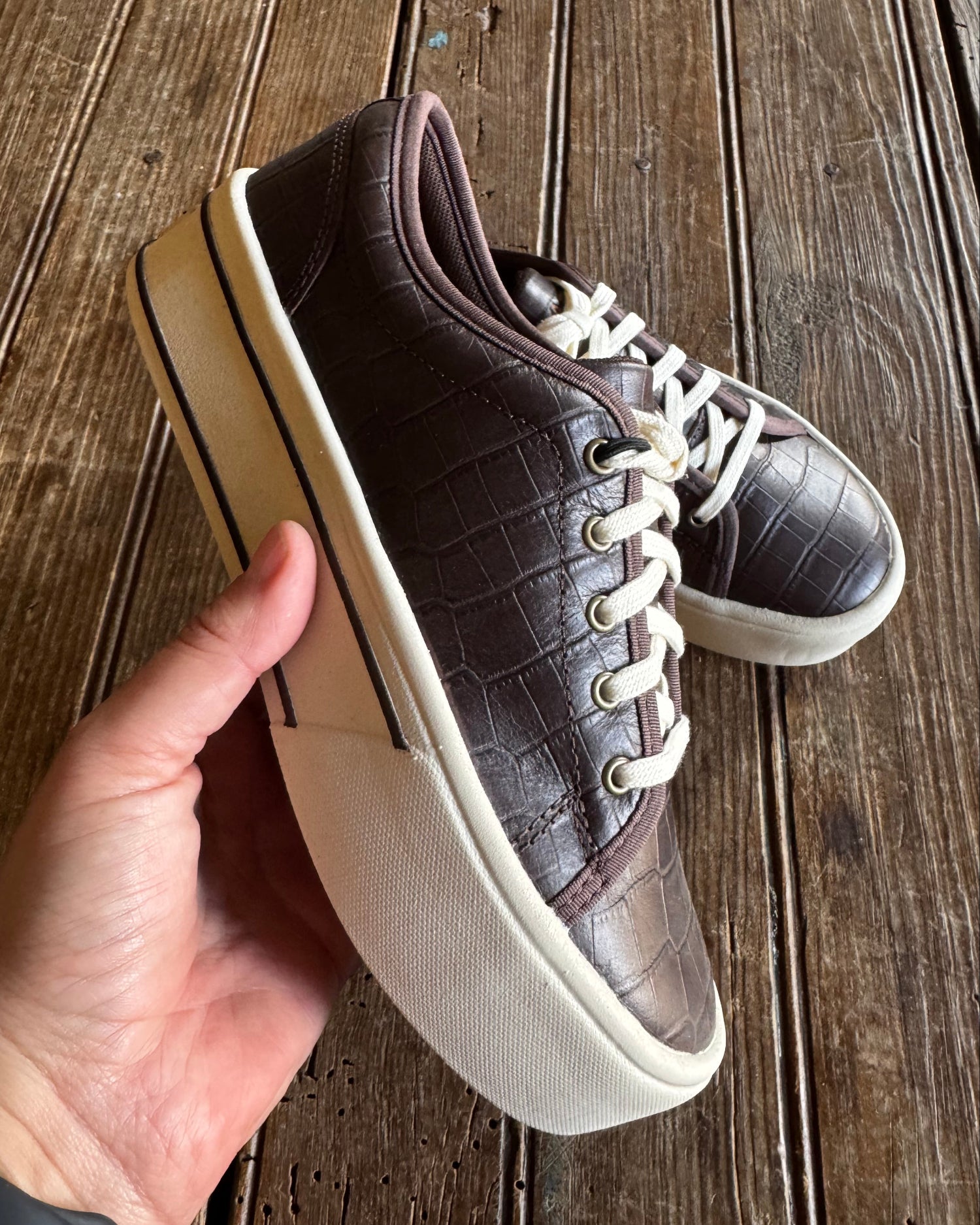 Brown crocodile-patterned sneaker with white sole held by a hand on a wooden surface